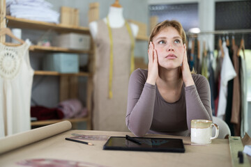 Exhausted from work, young female designer taking break in sewing studio, seated at table paper patterns in front of tablet with unfinished sketches and mug of coffee..