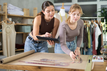 Two positive female dressmakers drawing dress design using paper and tablet in sewing workshop
