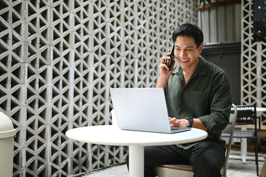 A man is sitting at a table with a laptop and talking on a cell phone. He is smiling while talking on the phone.