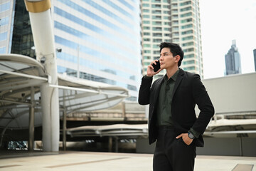 A man in a suit is talking on his cell phone while standing outside. He is in a good mood and is...