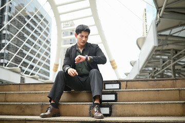 A man wearing a suit and tie is sitting on a set of steps. He is wearing a watch and he is checking the time.