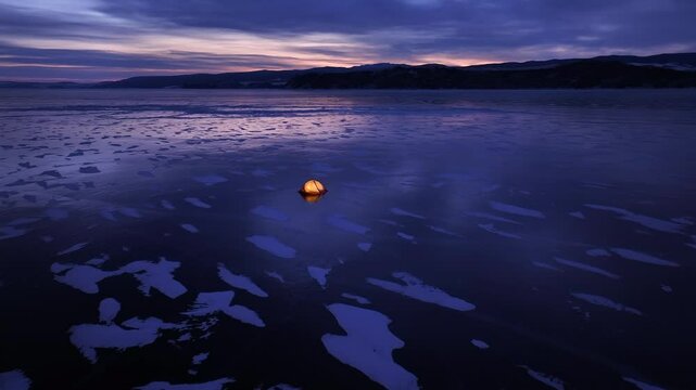 Illuminated orange tent on the icy reflecting surface of frozen lake at dusk