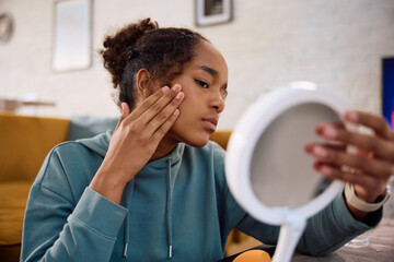 Black teenage girl examining skin on her face while looking herself in mirror.