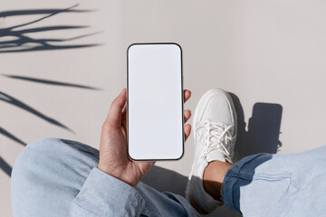 Person holding a smartphone with blank screen while sitting cross-legged in casual jeans and sneakers. Great for app mockups, tech branding, or lifestyle promotions.
