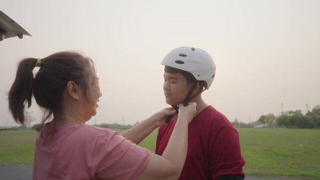 Asian mother putting helmet on teenage son before skateboarding in an open field. A moment of care, safety awareness, and family bonding in active outdoor life.