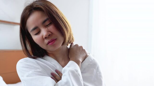 Asian woman sitting on bed in the morning, touching her neck and shoulder with a painful expression, feeling sore and stiff muscles in the upper back caused by sleeping in the wrong position at night