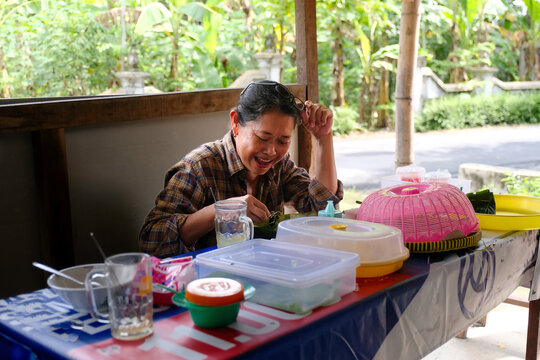 A smiling woman in a plaid shirt eats at an outdoor table, surrounded by plastic food containers, cups, and tropical greenery in the background