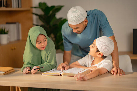 A Muslim dad sits with his son and daughter at home. They read books and do homework together. Everyone is happy and smiling.
