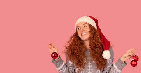 Festive woman wearing a Santa hat and holding Christmas ornaments in a cheerful pose against a pink background during the holiday season
