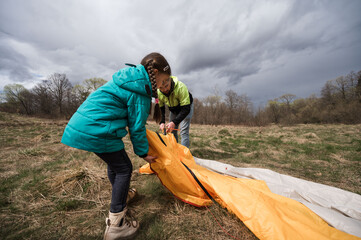 Young adventurers working together to set up a bright orange tent in a beautiful green field under...