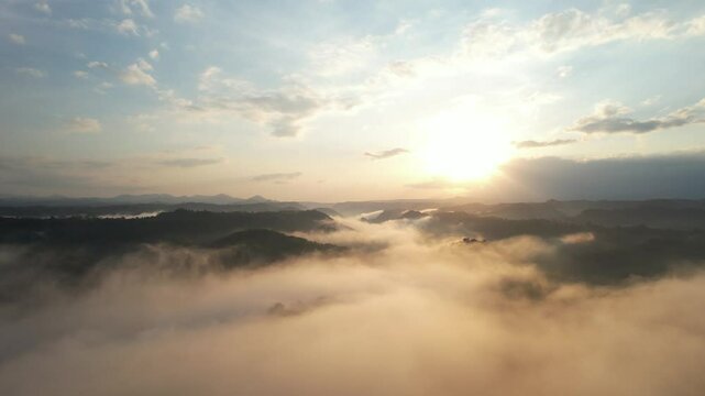 Aerial view of misty green hills during sunrise. Dense morning fog drifts over a lush tropical forest with golden sunlight and dramatic clouds. Perfect for nature, travel, or meditation themes.