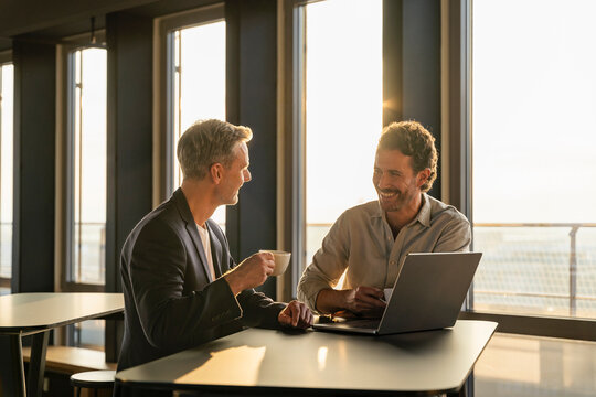 Two men having a business meeting in an office at sunset with coffee and a laptop