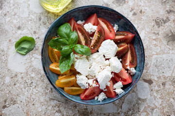 Tomato salad with ricotta and basil served in a blue bowl, horizontal shot on a light-brown granite background, top view