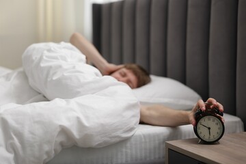 Sleepy man turning off alarm clock in bed indoors, selective focus