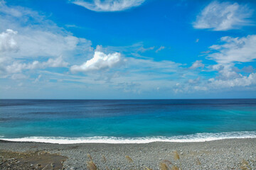 Vibrant Coastal Scene: Pebble Beach and Clear Blue Water. A tranquil view of small waves washing onto a pebble beach under a vivid blue sky with scattered white clouds.
