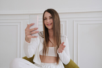A happy young woman smiles brightly while looking at her smartphone screen, clearly engaged with entertaining content. Shot in natural indoor lighting, this authentic moment represents modern digital 
