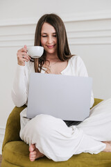 A female freelancer enjoys her coffee while working on a laptop in a comfortable armchair, demonstrating a productive yet relaxed work-from-home setup. Warm natural lighting highlights her modern home