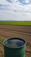 Obraz premium A Green Barrel of Water in a Dried-up Rice Paddy Field Under a Partly Cloudy Sky