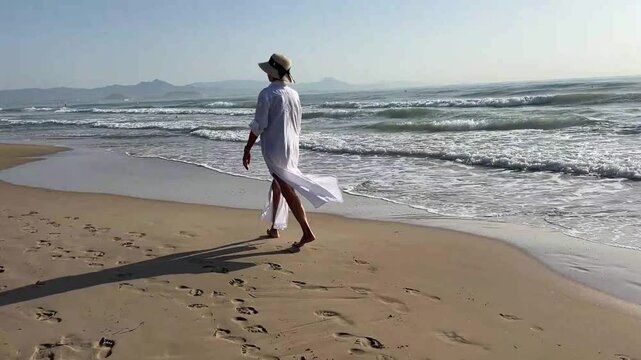 Senior woman in her 80s strolling along the beach at sunrise, wearing a white caftan and wide-brimmed hat, surrounded by calm sea and warm morning light
