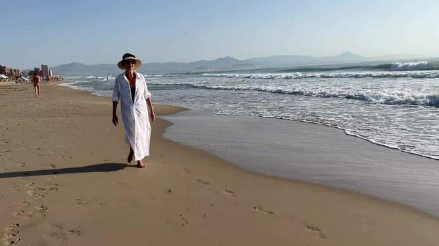 Elderly woman in her 80s walking peacefully along the beach wearing a white caftan and sun hat, enjoying the coastal breeze and morning light in a serene seaside setting
