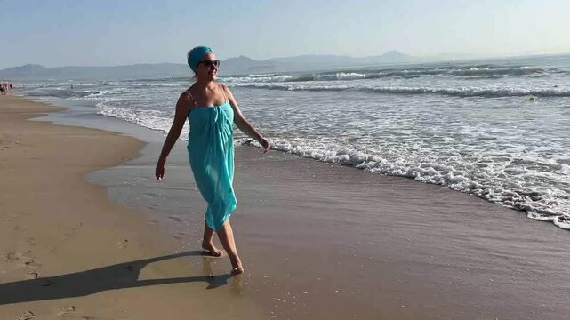 Middle-aged woman wearing a blue pareo walking along the shoreline, enjoying the gentle waves and sunshine on a peaceful beach day