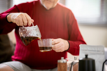An elderly man is happily making coffee on a bright morning at his home.
