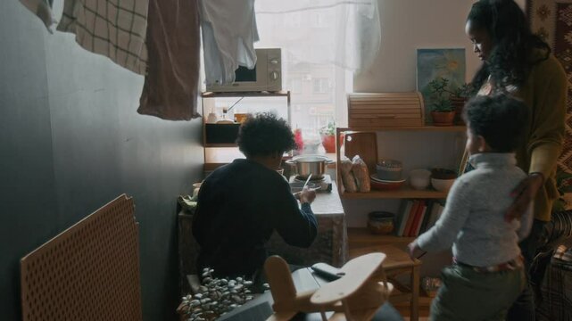 Poor African American mother preparing dinner for two of her sons, sitting in kitchen area of small lived-in apartment