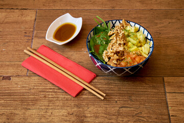 A bowl of food with a red napkin and chopsticks on a wooden table. The bowl contains a variety of food items such as vegetables, nuts, and fruit