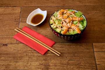 A bowl of food with shrimp and avocado on top. A red napkin with chopsticks on it