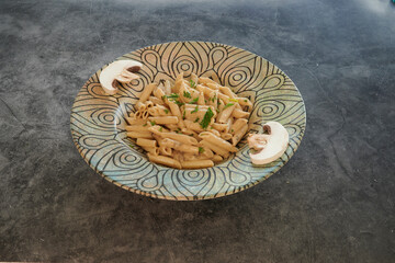 A bowl of pasta with mushrooms on top is placed on a counter. The mushrooms are positioned on the edge of the bowl, while the pasta is in the center. The bowl is a blue and white design
