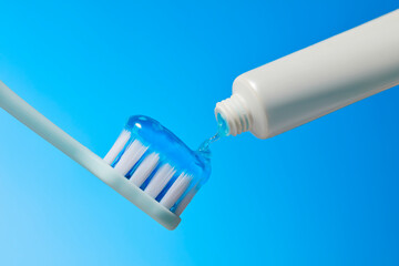 A blue toothbrush is being filled with white toothpaste from a white tube. The background is light...
