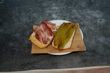 A sandwich with ham and peppers on a plate. The plate is on a table. The sandwich is cut in half