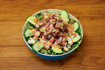 A bowl of food with a lot of vegetables and a piece of meat. The bowl is blue and is on a wooden table