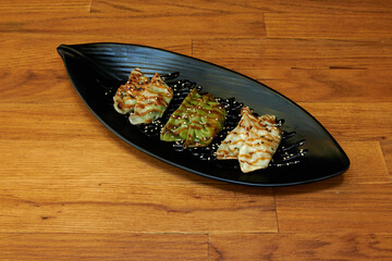 A black plate with four dumplings on it. The plate is on a wooden table. The dumplings are different colors and have different sauces