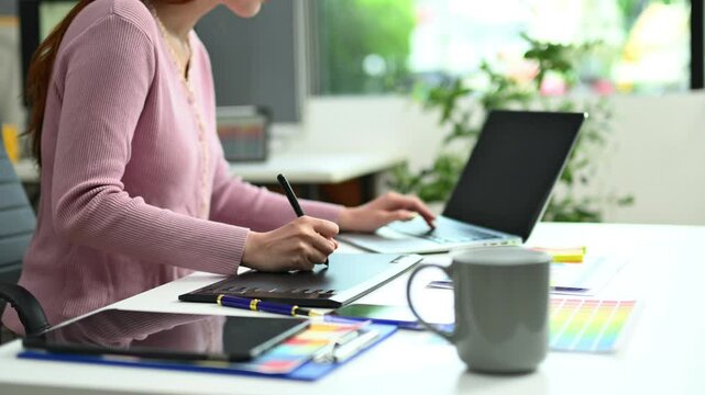 Young graphic designer using pen tablet and laptop at creative desk in modern office. Surrounded by color palettes and design tools.