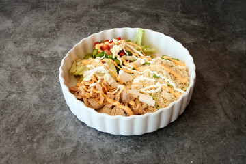 A white bowl with a variety of food in it. The bowl contains a salad, rice, and meat
