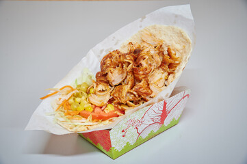 A wrap with chicken and vegetables is sitting in a paper box. The wrap is covered in a sauce and has a lot of toppings
