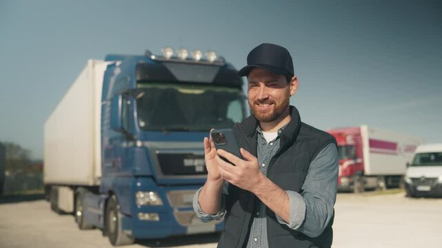 Smiling bearded Caucasian male wearing dark cap and padded vest standing outdoors near blue truck. Holding smartphone and watching video or reading messages. Enjoying break before next delivery.