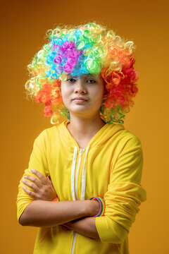 Smiling Asian teenage girl wearing rainbow LGBTQ flag wig standing with arms crossed on yellow background