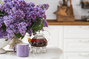 A large purple bouquet of lilacs, a teapot and a cup of tea on the table. In the background is the...