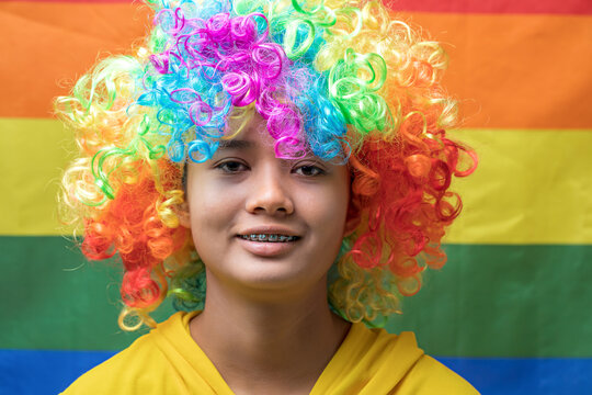 Smiling Asian teenage girl with braces in rainbow lgbtq flag wig looking at camera with rainbow flag in background, close up