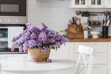 A large purple bouquet of lilacs on the table in a wicker basket. In the background is the interior of a modern white kitchen. The concept of home comfort and celebration.