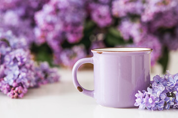 A lilac cup of tea or coffee on the table. In the background is a large purple bouquet of lilacs. The concept of home comfort and drinks.