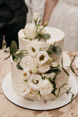 white wedding cake decorated with white large roses and green leaves stands on a white plate in the background of the bride and groom on their wedding day
