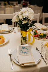 fresh white flowers in a vase wrapped in a yellow ribbon stand on a served table at a wedding, in a plate on a napkin is an invitation card with the name of the guest