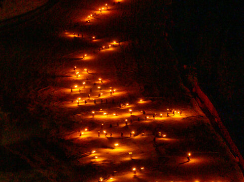 A high-angle shot shows a torchlight procession snaking down a snowy slope at night. Many small, bright lights illuminate the path. Monte Santo di Lussari, Tarvisio, Friuli Venezia Giulia, Italy.