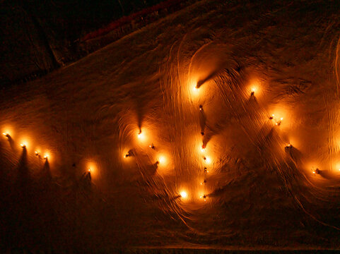 A high-angle shot shows a torchlight procession snaking down a snowy slope at night. Many small, bright lights illuminate the path. Monte Santo di Lussari, Tarvisio, Friuli Venezia Giulia, Italy.