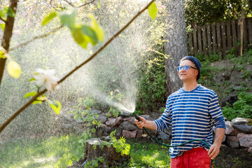 Middle-aged man in a sun hat watering lawn and garden plants with a hose in the backyard on a sunny...