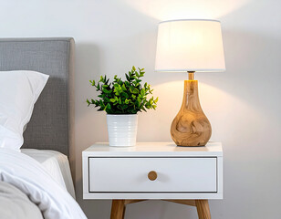 White nightstand with wooden table lamp and green potted plant against neutral background. Modern bedroom detail with gray headboard and white bedding.