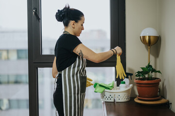 A woman wearing an apron prepares cleaning supplies near a window in a bright interior. She is...